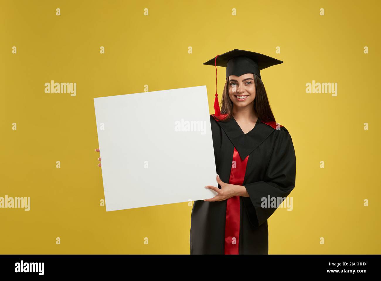 Front view of girl in mortarboard standing, smiling, looking at camera. Pretty young female graduating from college, holding, showing. Isolated on yellow studio mockup background. Stock Photo