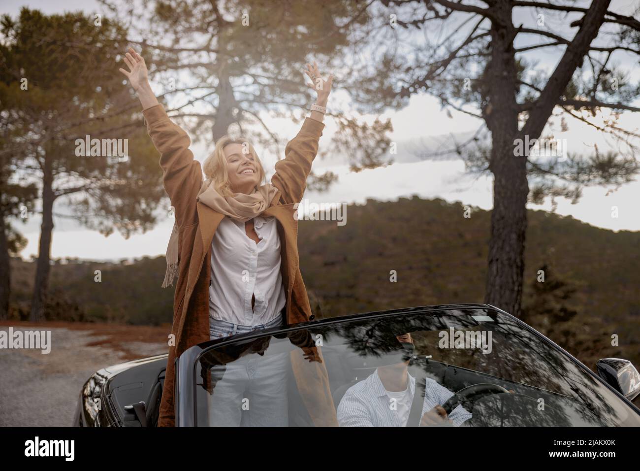 Pretty young smiled woman stands on front seat in convertible car with her hands raised up Stock Photo