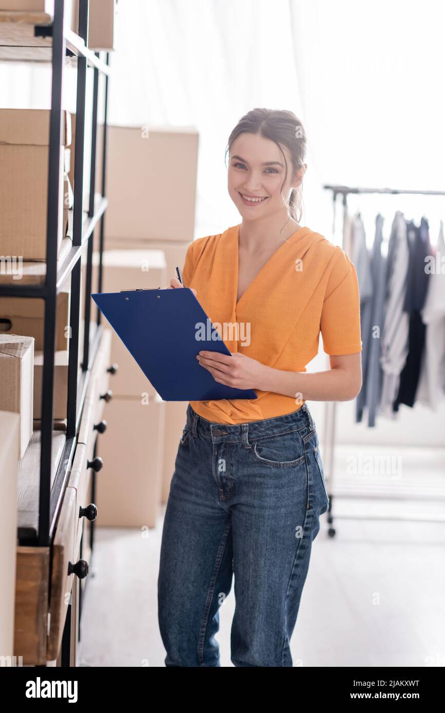 Cheerful owner of online web store holding clipboard and looking at camera near boxes on rack Stock Photo
