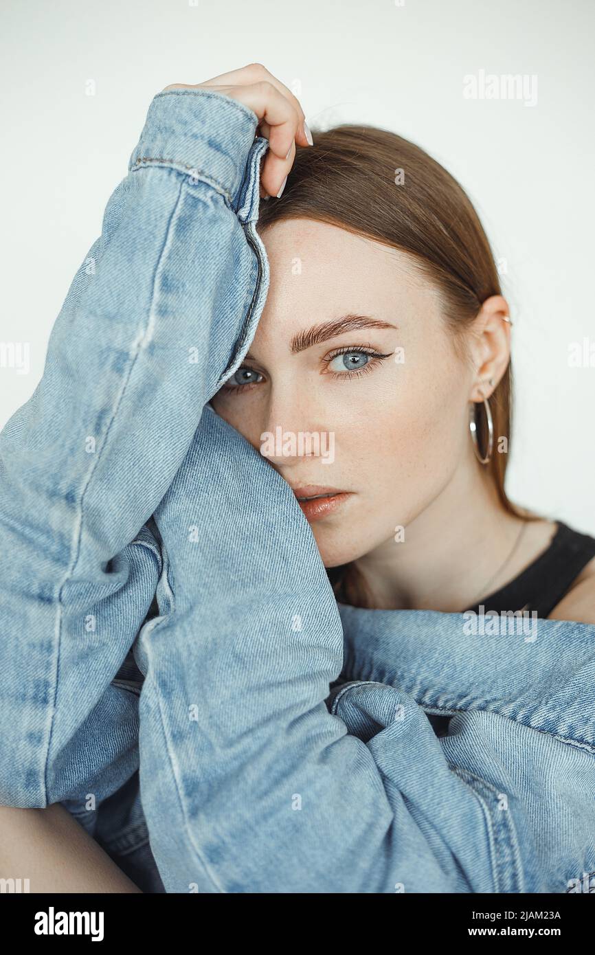 Vertical of calm, serious dark-haired woman looking at camera, wearing denim jeans jacket. Hugging herself. Close-up Stock Photo
