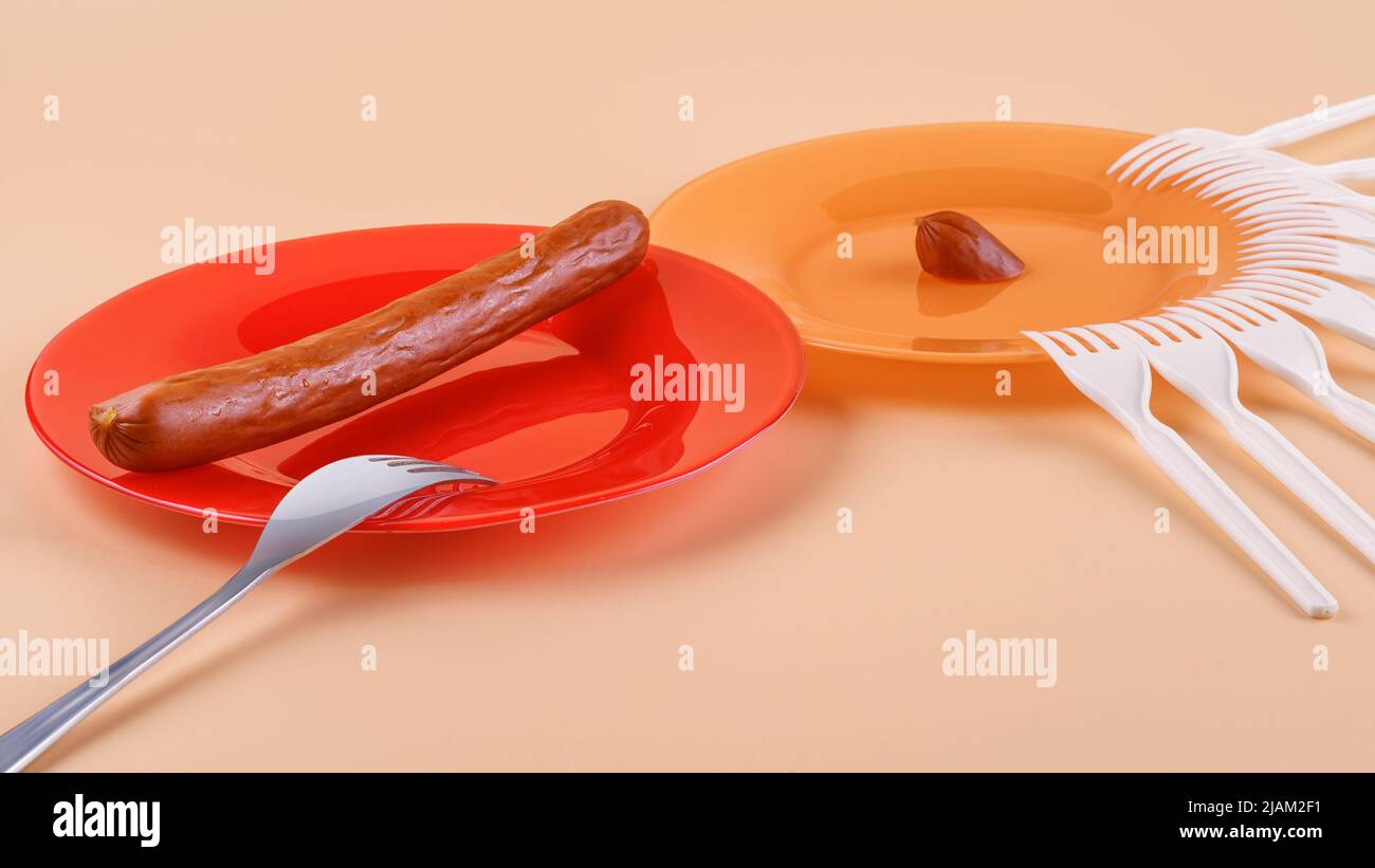 Two plates, two different sausage portions and many forks. Metaphor for disproportion and inequality in allocating budget, wages, resources, funds, fi Stock Photo