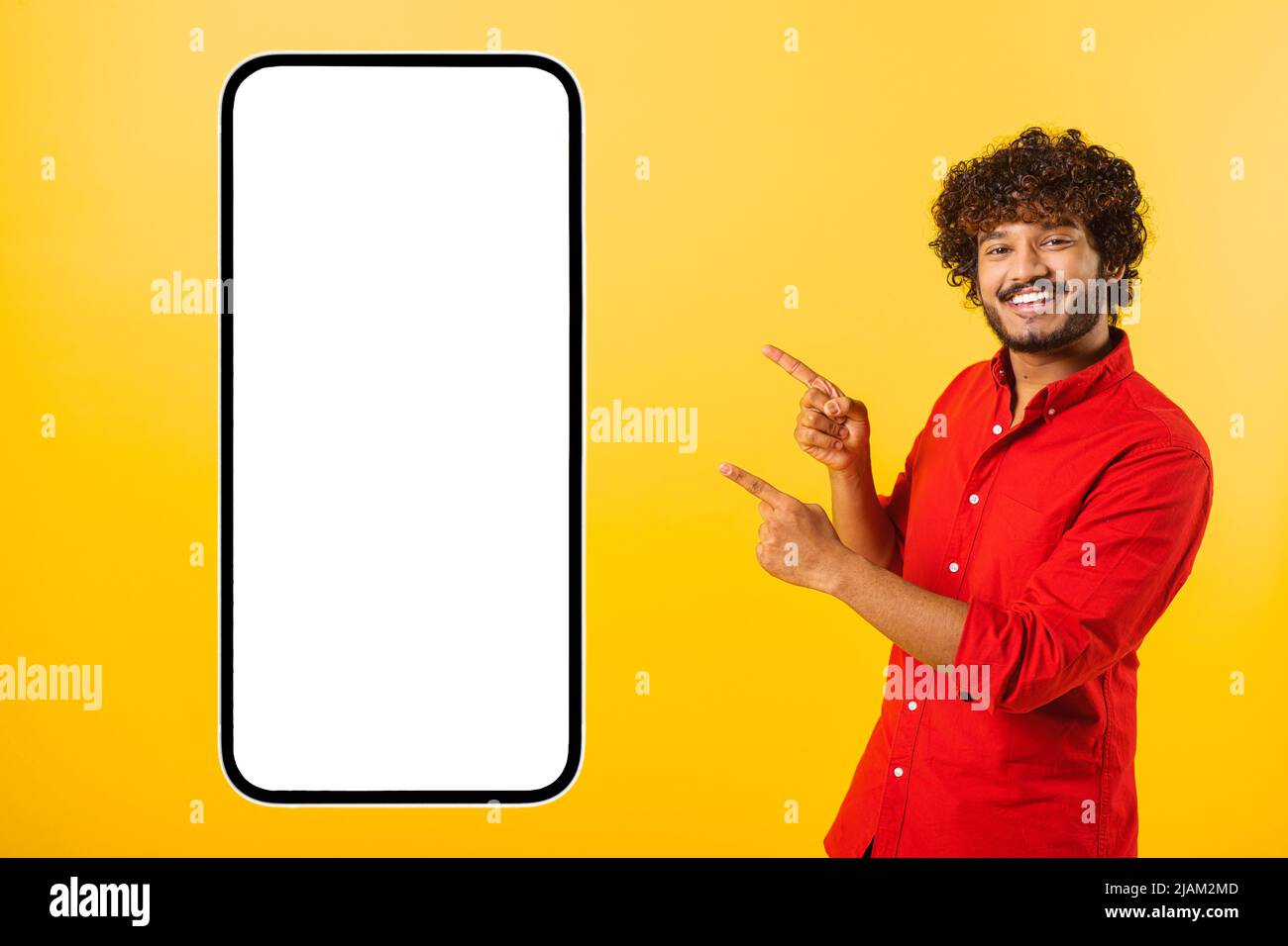 Playful handsome curly indian bearded man points fingers at huge smartphone with empty blank screen isolated on yellow, happy latin guy presenting mobile app, showing best deal, copy space, template Stock Photo