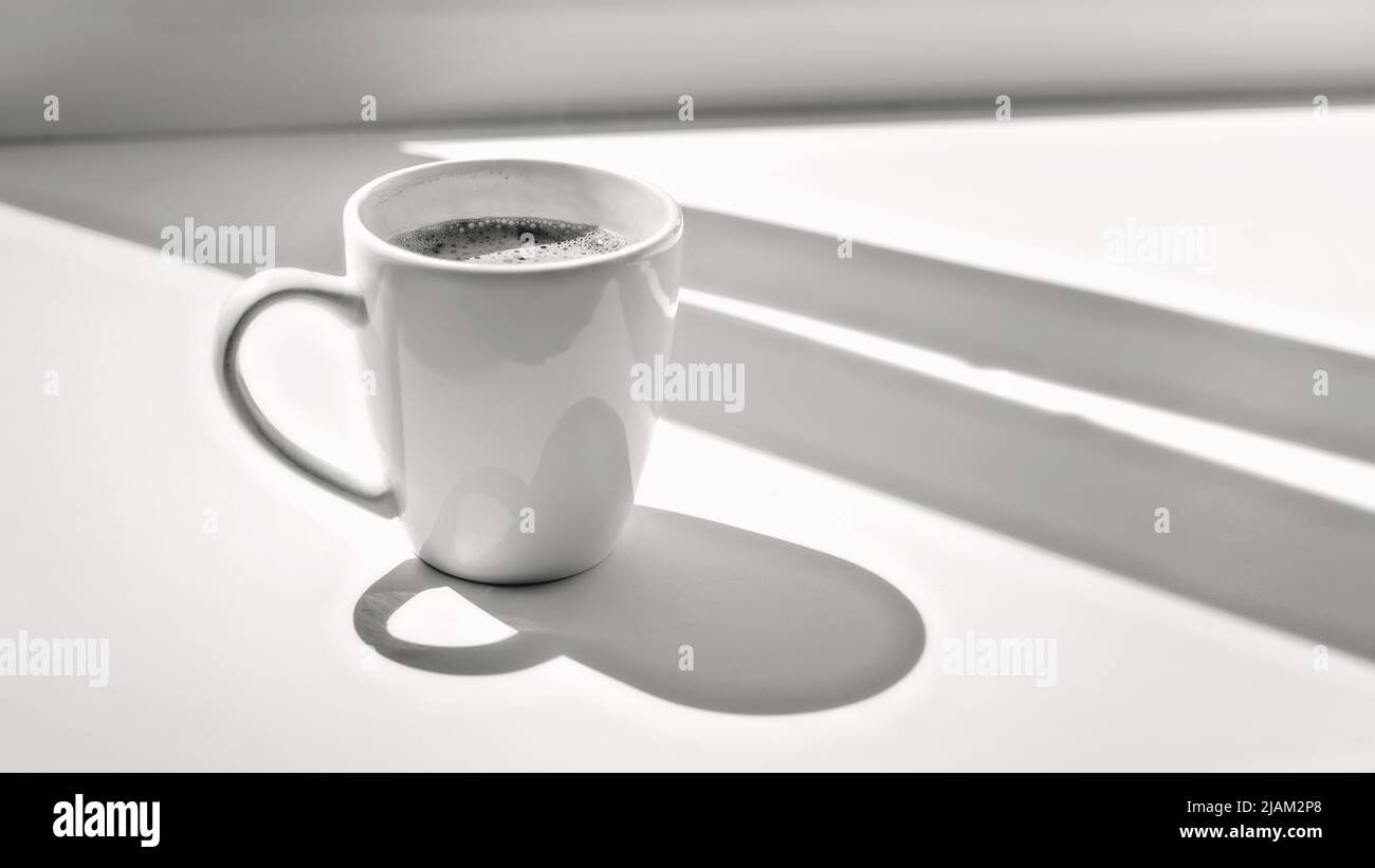 White cup of coffee on a sunlit window sill. Black and white image, selective focus Stock Photo