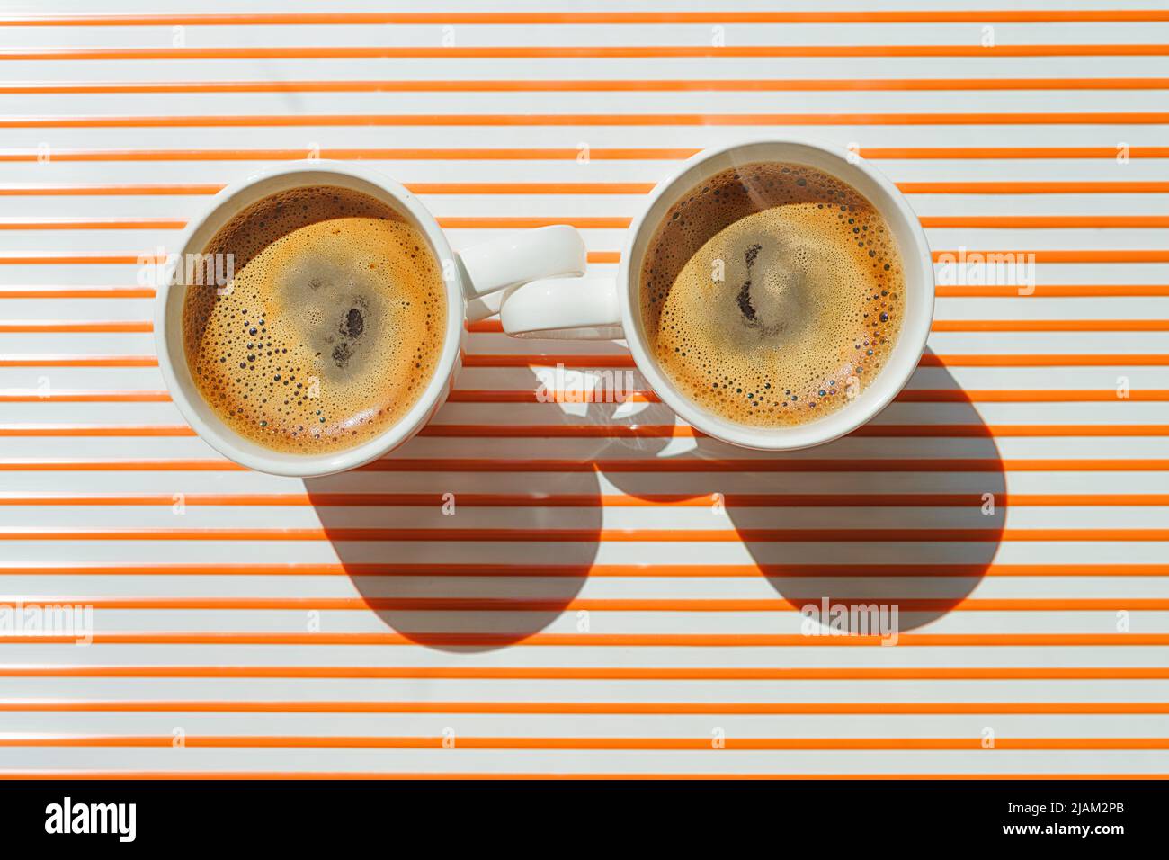 Two white coffee cup close one to other on orange striped table at direct sunlight. Top view with shadow Stock Photo