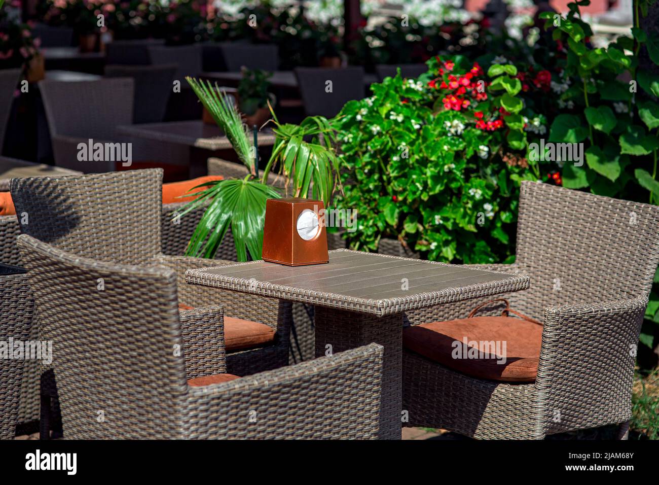 wicker rattan plastic fittings on terrace of street restaurant lit by sun, square table with napkin holder and chairs with soft cushion nearby green p Stock Photo