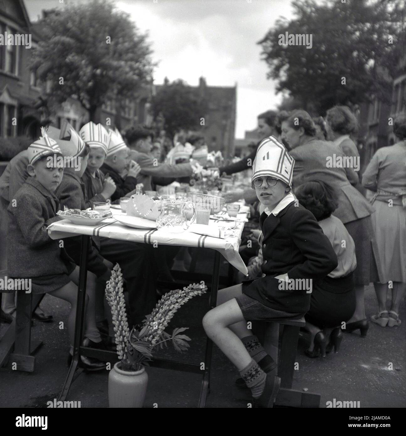 1953, historical, street party celebrating the Coronation of Queen Elizabeth II to the throne, a group of people sitting at table in a street, with young boys at the end of the table, sitting wearing hats, some with the letter E on, England, UK. Stock Photo