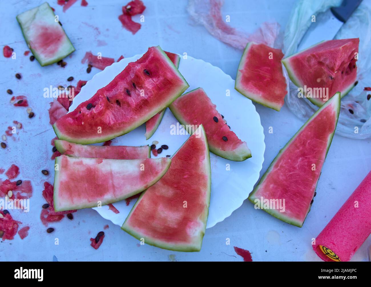 portions of watermelon eaten by children messed up on a plate and a white table. All dirty with juice and watermelon seeds outdoors. portions of water Stock Photo