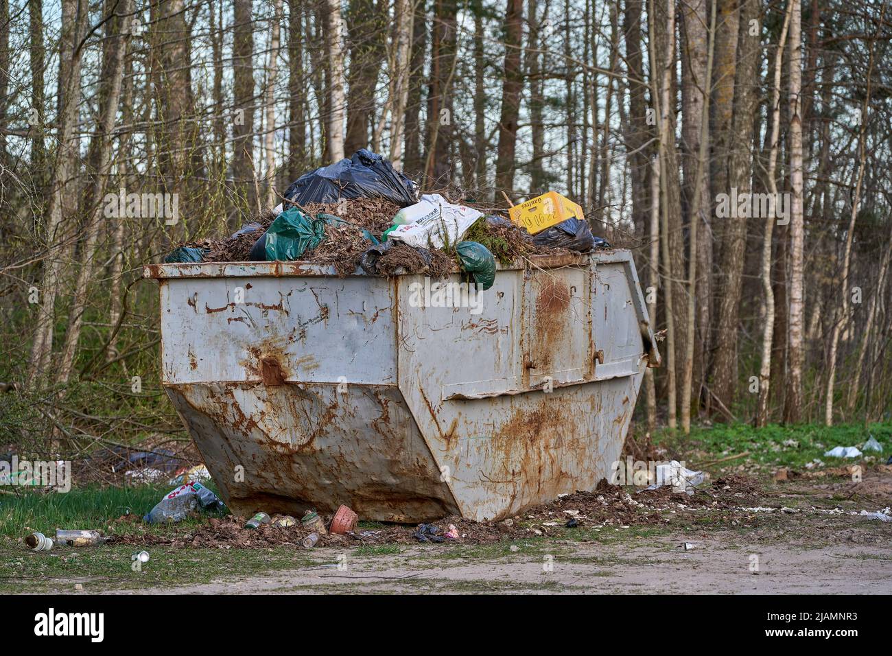 An old rusty metal container overflowing with rubbish. Some garbage lying around. Many trees at the background. Stock Photo