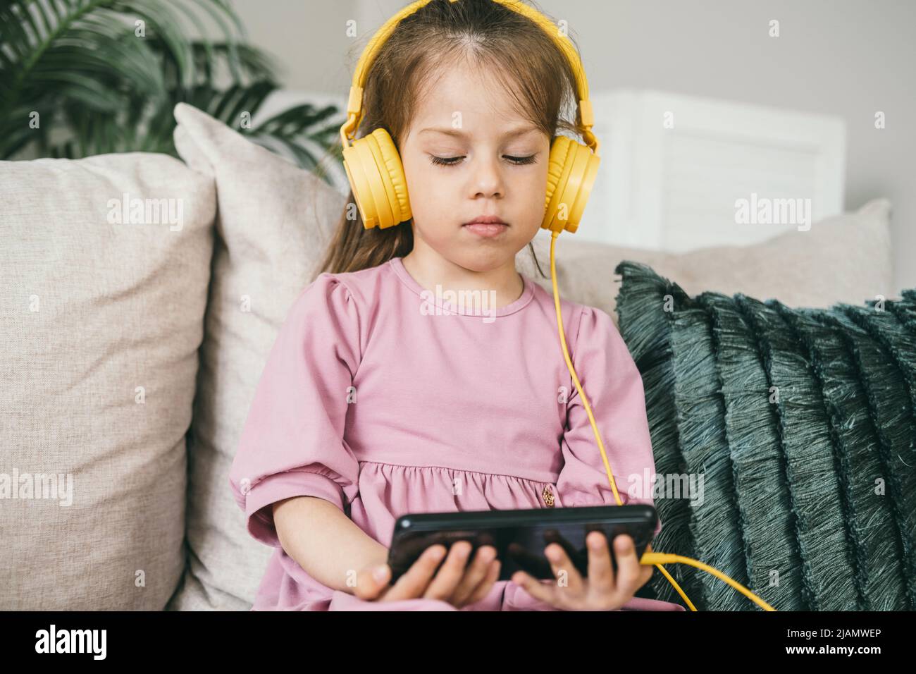 A little girl in yellow headphones sits on a sofa with a phone Stock ...