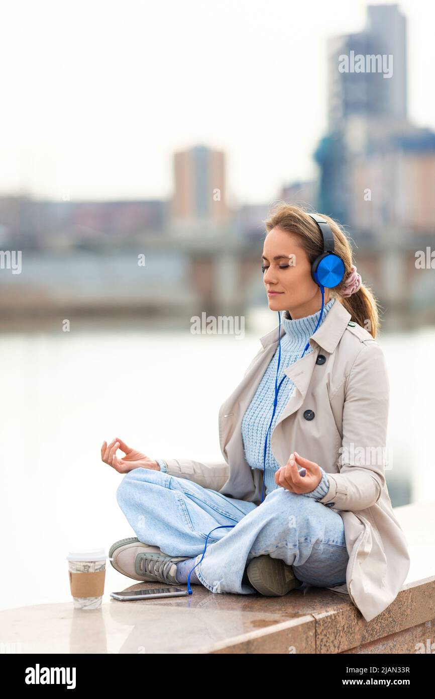 A young woman meditates on the embankment - she sitting in headphones and listening to relaxing music. Rest from office bustle and city noise. Vertica Stock Photo