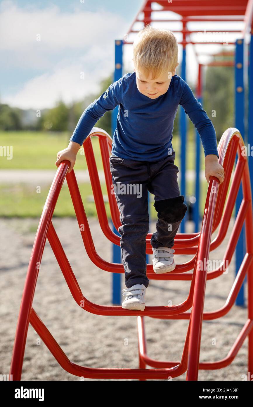Brave small child playing and climbing arch ladder in public kid playground during beautiful day in summer time. Stock Photo