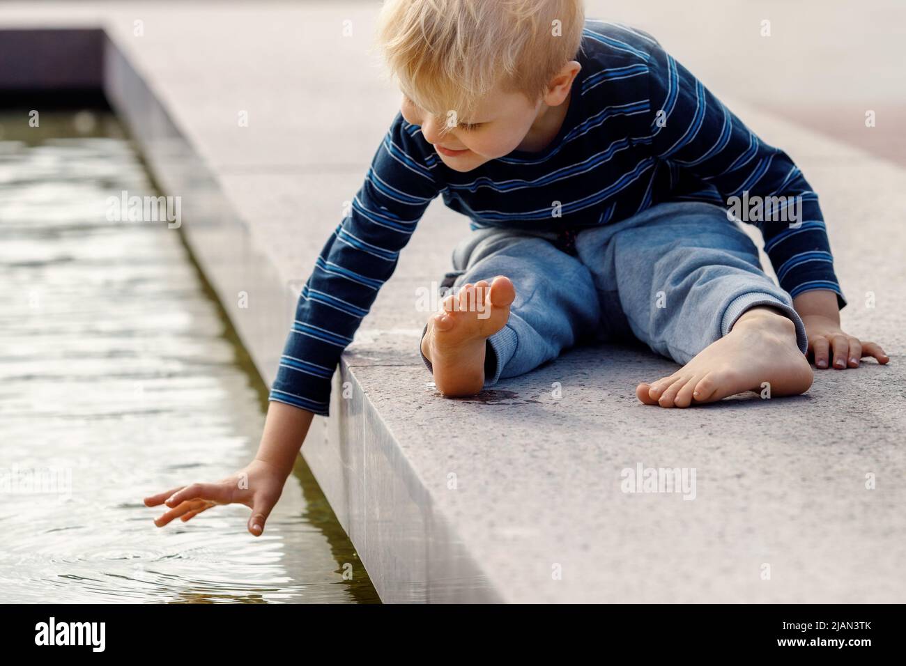 A little boy in the city plays near the fountain with water at summer. Happy boy touching water in fountain. Stock Photo