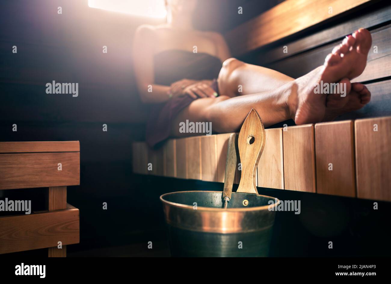 Sauna, steam room bath. Woman relaxing in spa. Wellness and warm temperature therapy in dark wood home in Finland. Water bucket and ladle. Stock Photo