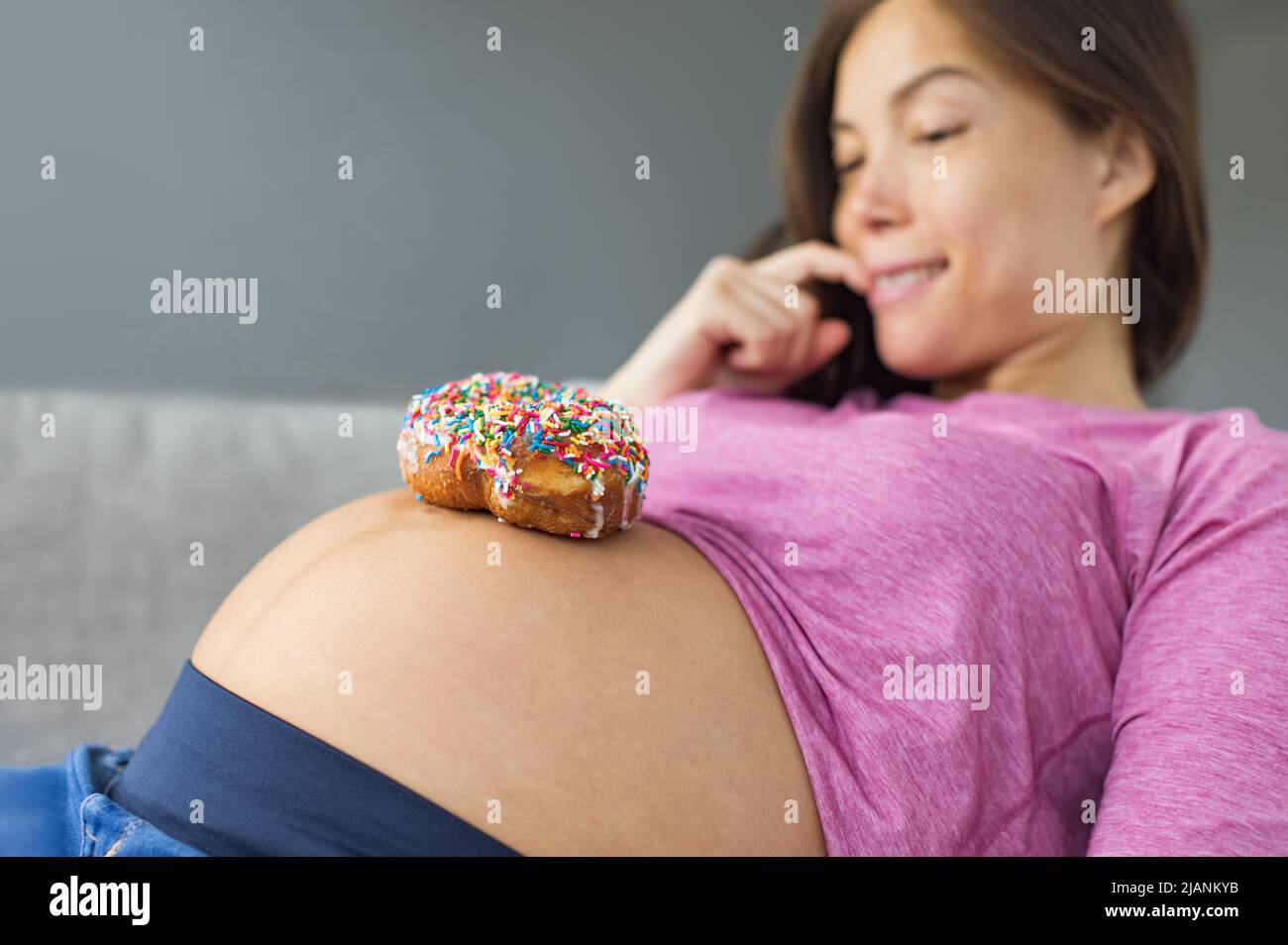 Unhealthy food during pregnancy. Sugar temptation craving Asian pregnant woman trying to resist eating a sweet donut on her baby belly. Funny eating Stock Photo