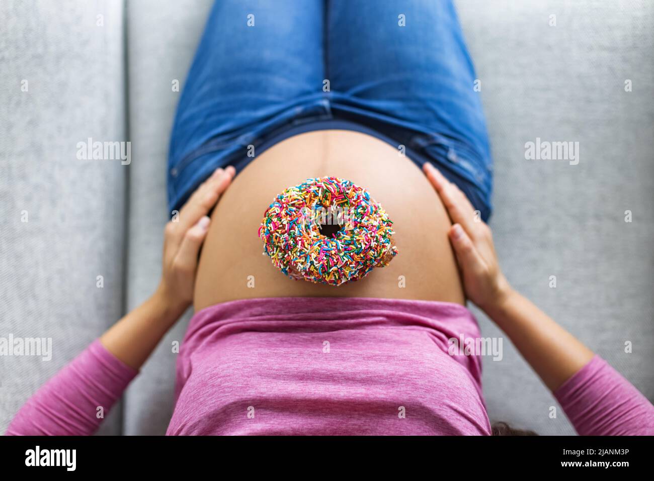 Pregnant woman with donut on belly top view. Cravings of desserts and sweets during pregnancy, Pastry with birthday cake sprinkles on baby bump for Stock Photo
