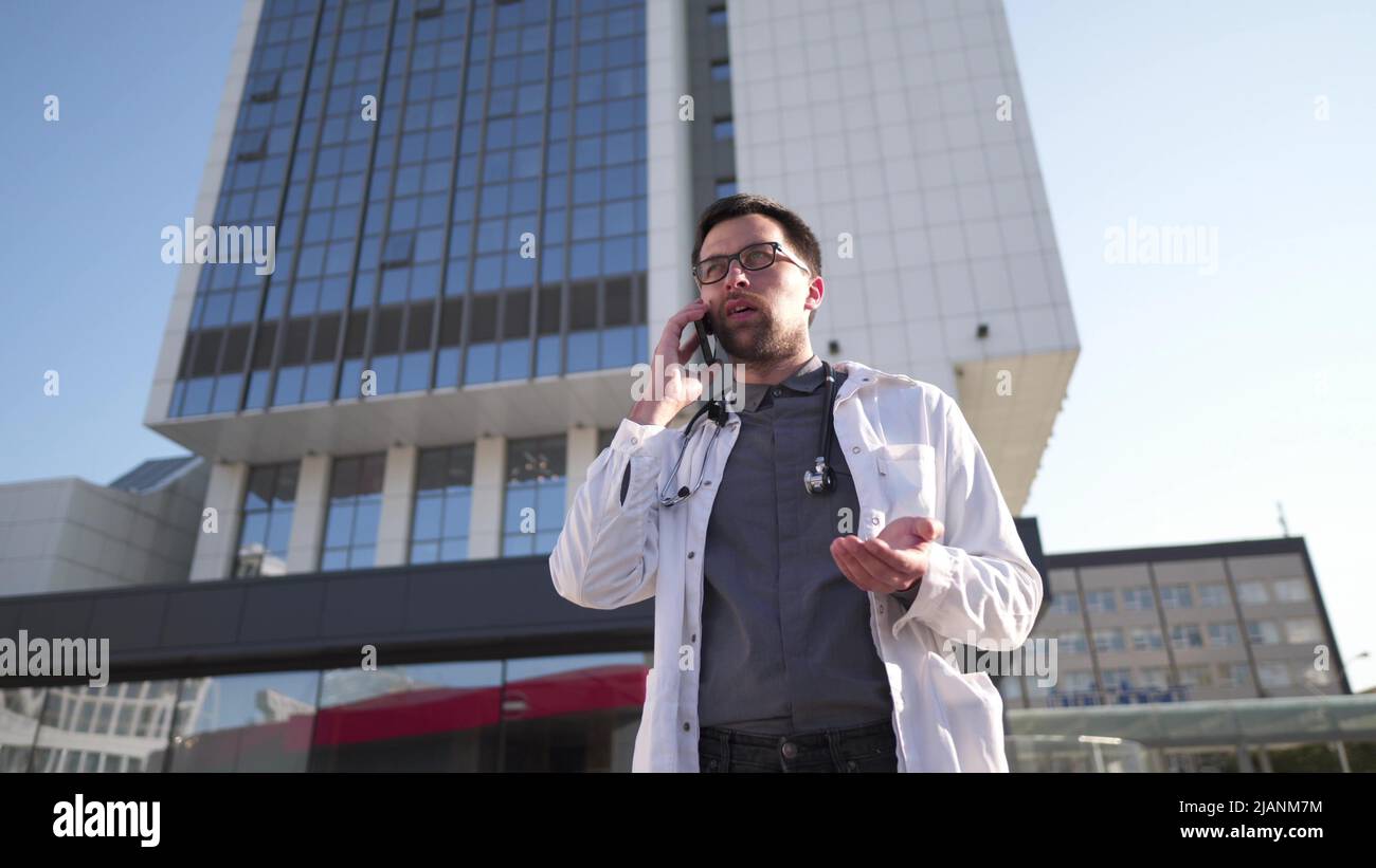 Young Caucasian doctor in white medical coat consults patient on cell phone during break outside hospital. Medical worker talking on mobile phone near Stock Photo