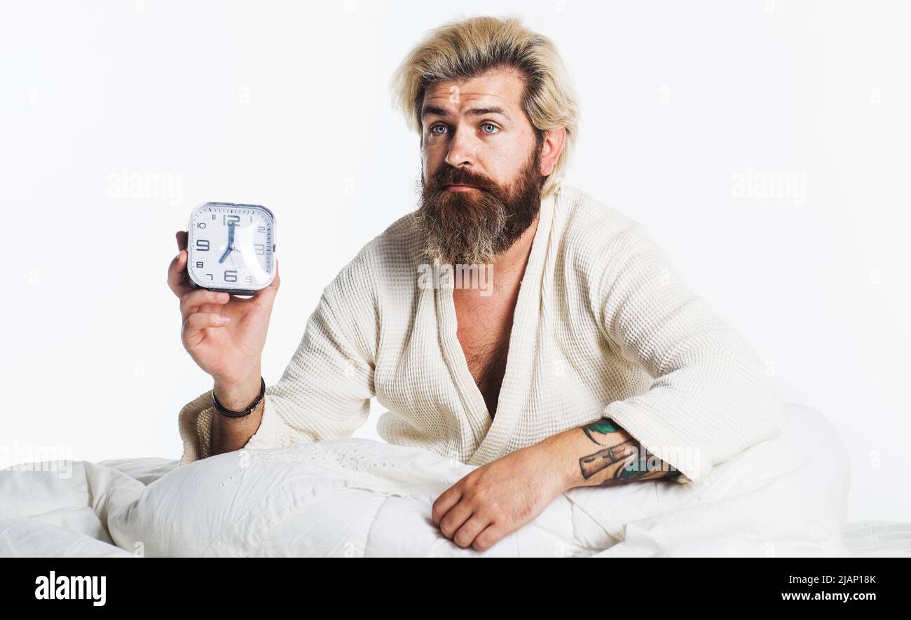 Sleepy bearded man with alarm clock in bed. Morning time. Awaking. Stock Photo