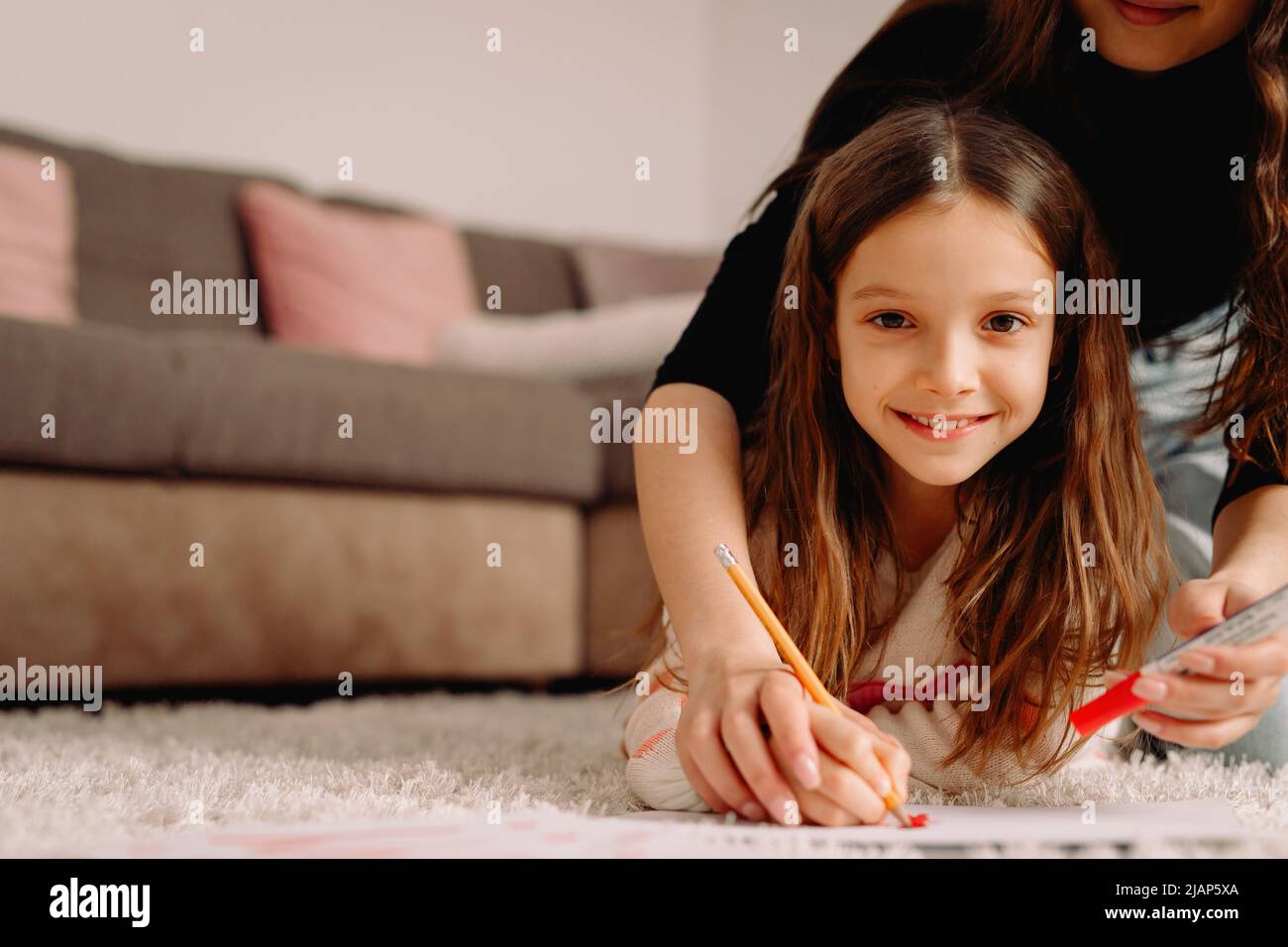 Little pretty blond girl smiling together with her mother at home drawing on the papre using red pencil, family time together concept. Stock Photo