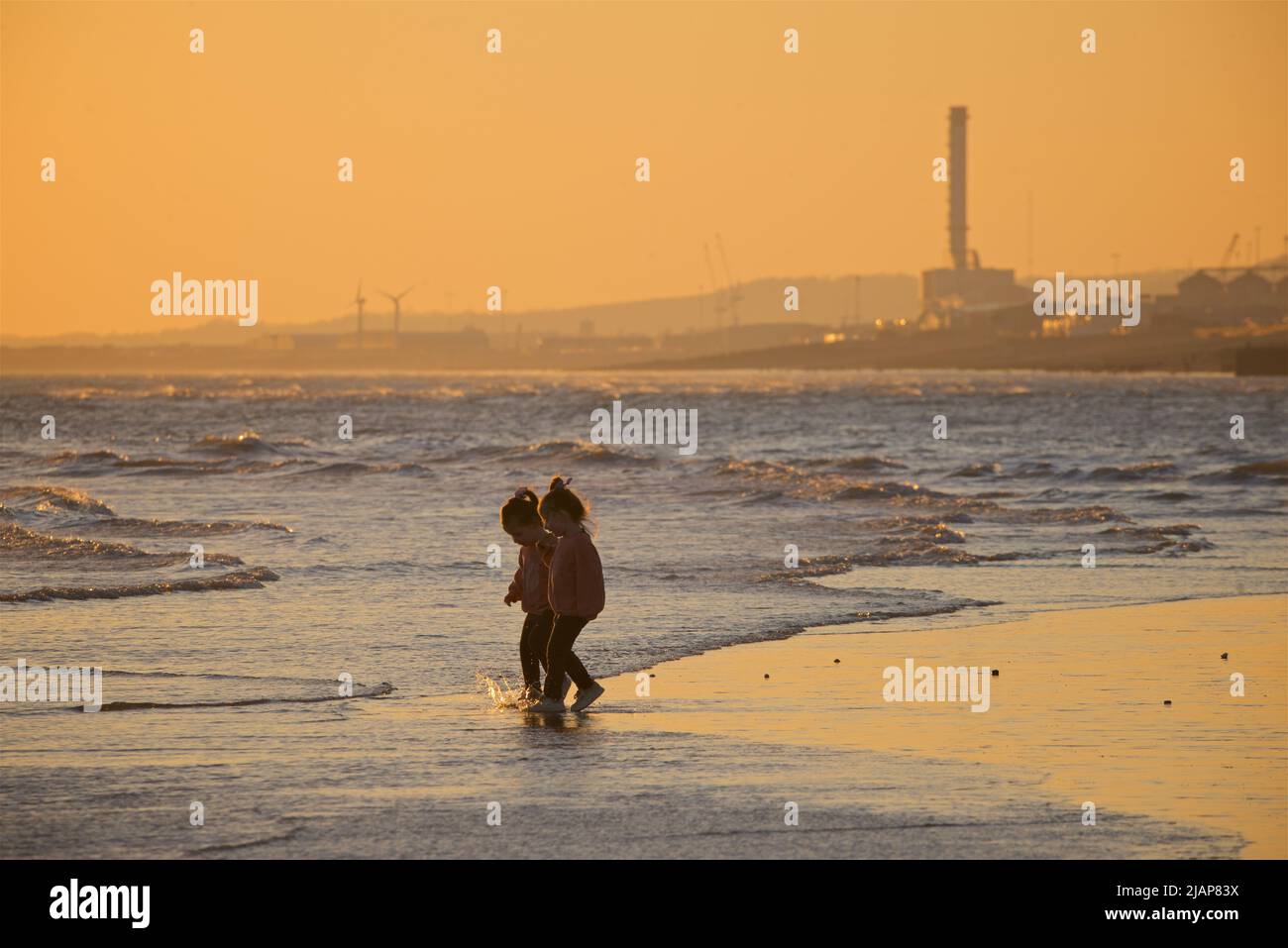 Two young girls walking along the shoreline at low tide, Brighton, Sussex, England, UK. Sunset / dusk. Shoreham Power Station in the background. Stock Photo