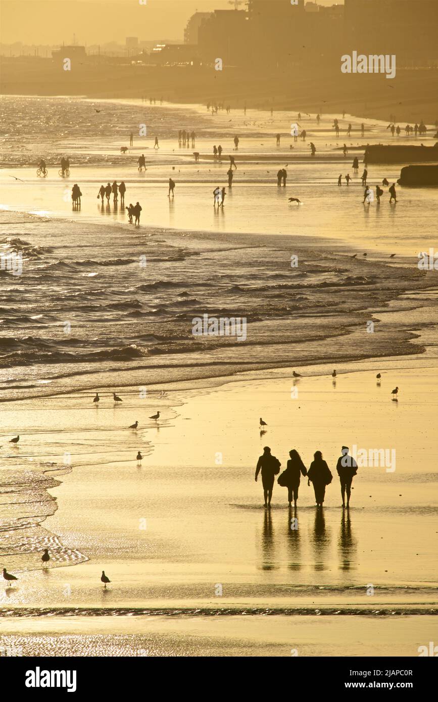 Silhouetted shapes of friends together and other people on the beach at low tide, Brighton & Hove, East Sussex, England, UK. {hotographed from the Palace Pier at sunset Stock Photo
