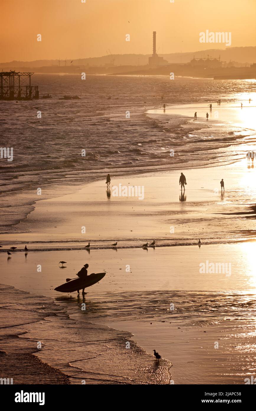Silhouetted shapes of man with a kayak on the beach at low tide, Brighton & Hove, East Sussex, England, UK. Photographed from the Palace Pier with the remains of the West Pier, and Shoreham Power Station in the distance. Stock Photo