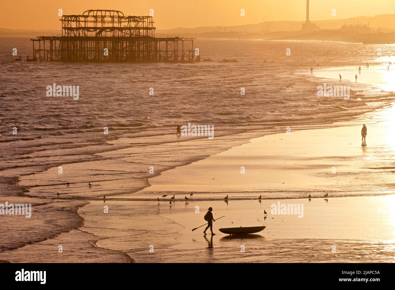 Silhouetted shapes of man with a kayak on the beach at low tide, Brighton & Hove, East Sussex, England, UK. Photographed from the Palace Pier with the remains of the West Pier, and Shoreham Power Station in the distance. Walking in from the sea Stock Photo