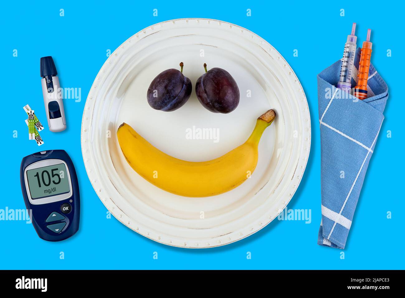 Banana and plum in the form of a smiling emoticon on a white plate and pen for insulin syringes and a blood glucose meter as cutlery on a blue table. Stock Photo