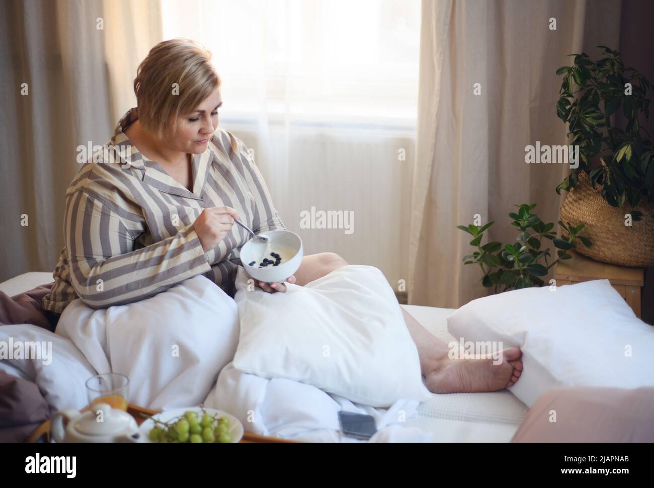 Happy overweight woman reading book and having breakfast in bed at home. Stock Photo