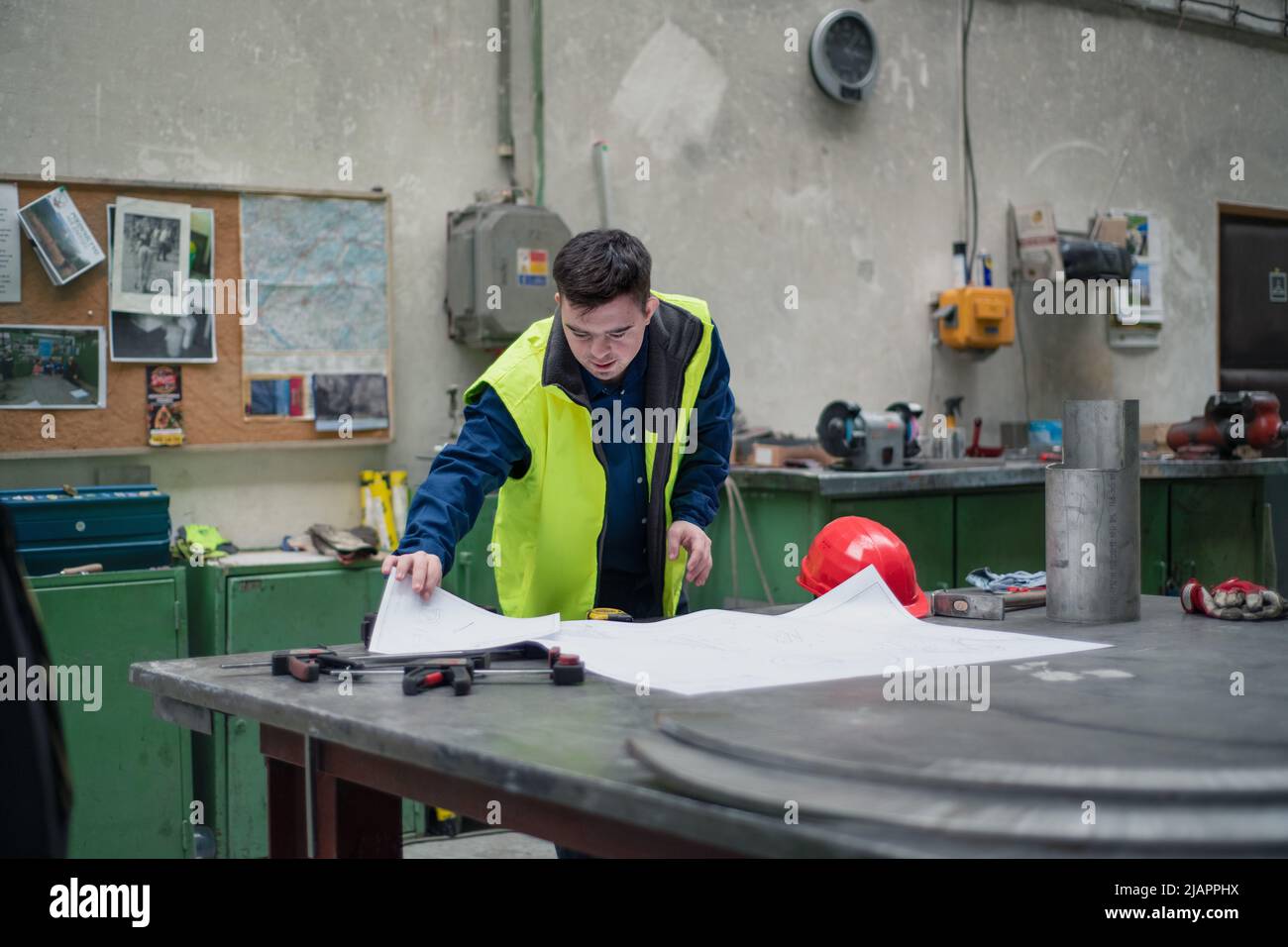 Young man with Down syndrome looking at blueprints when working in industrial factory, social integration concept. Stock Photo