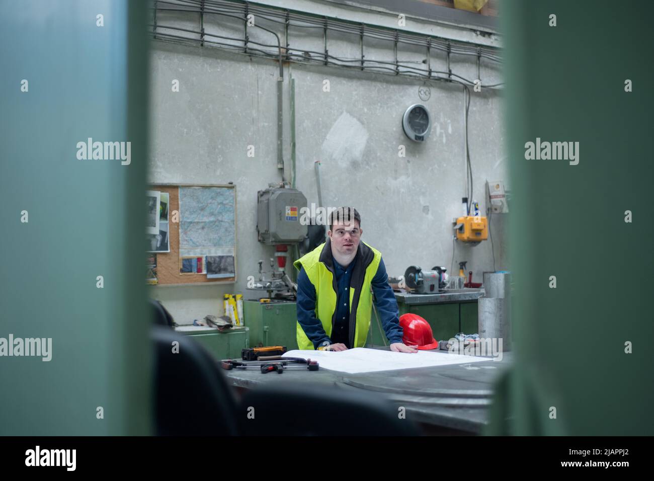 Young man with Down syndrome looking at blueprints when working in industrial factory, social integration concept. Stock Photo