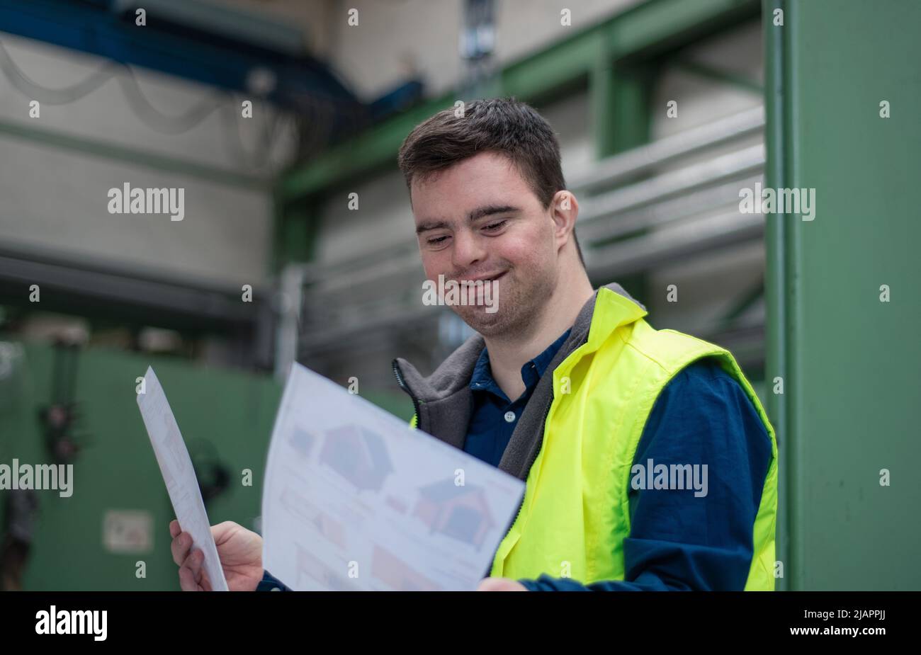 Young man with Down syndrome looking at blueprints when working in industrial factory, social integration concept. Stock Photo