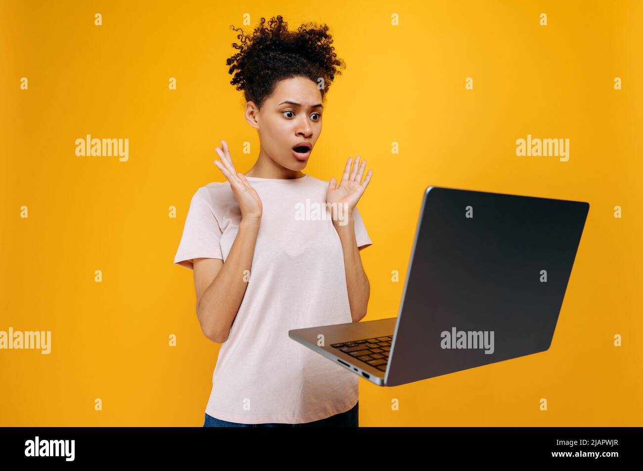 Stunned shocked african american girl, in a t-shirt, with curly hair, looks in surprise at the laptop screen, gesticulates with her hands, saw the unexpected news, stands on isolated orange background Stock Photo