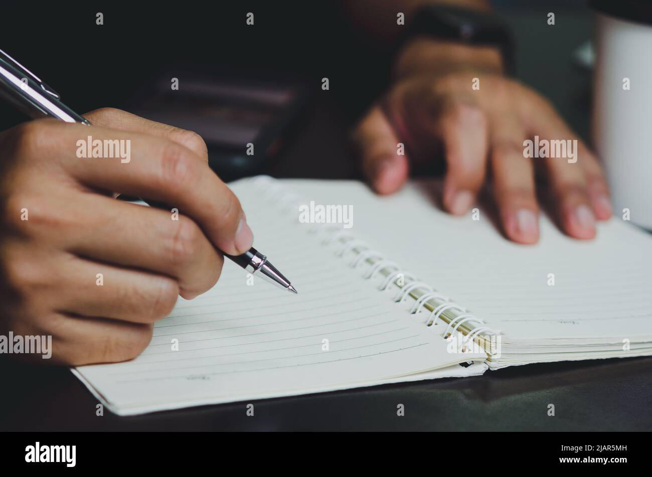 Man holding a pen and writing on a notebook on his desk Stock Photo - Alamy