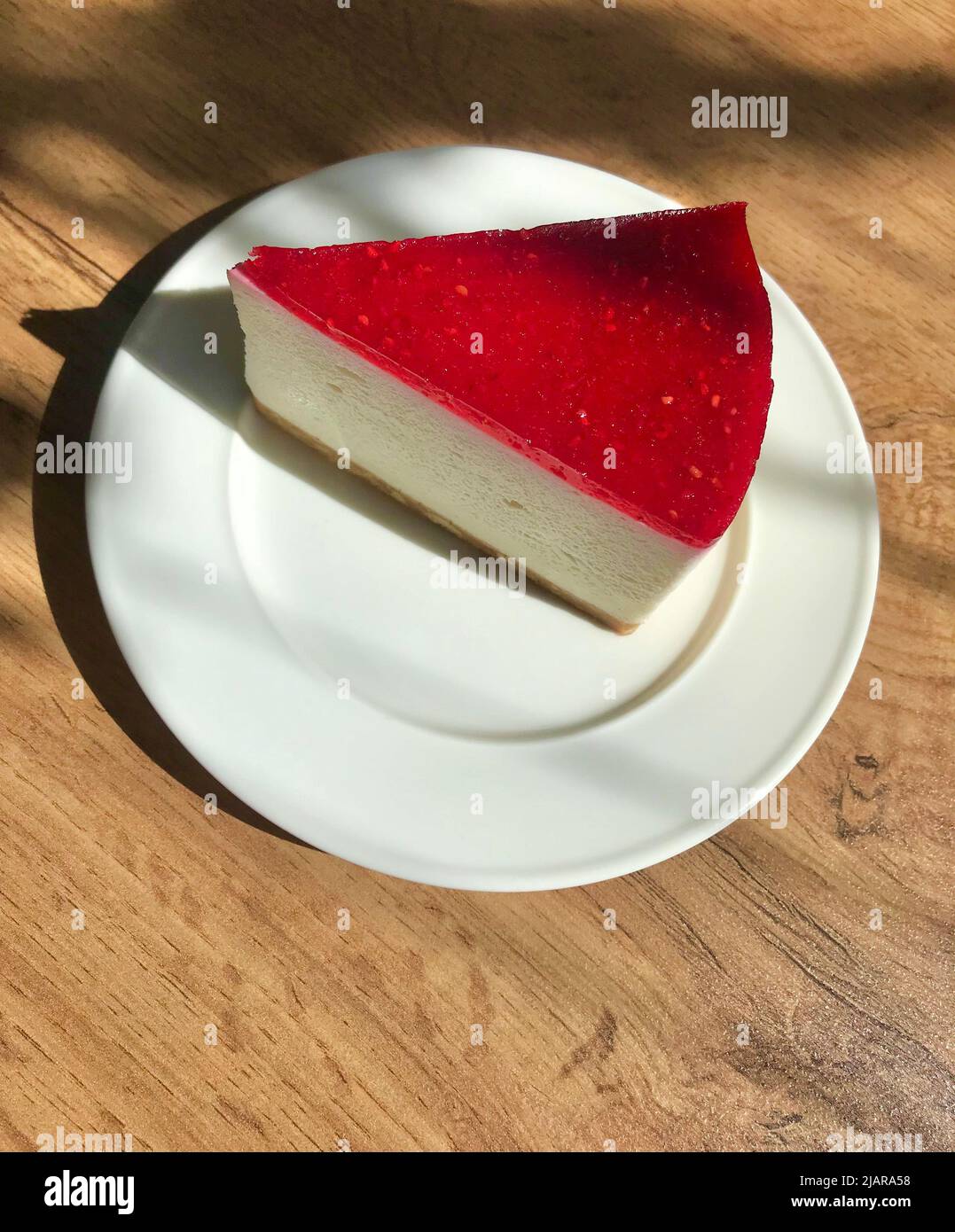 Raspberry cheesecake on a white plate. Wooden table with shadows. Breakfast in a cafe. Stock Photo