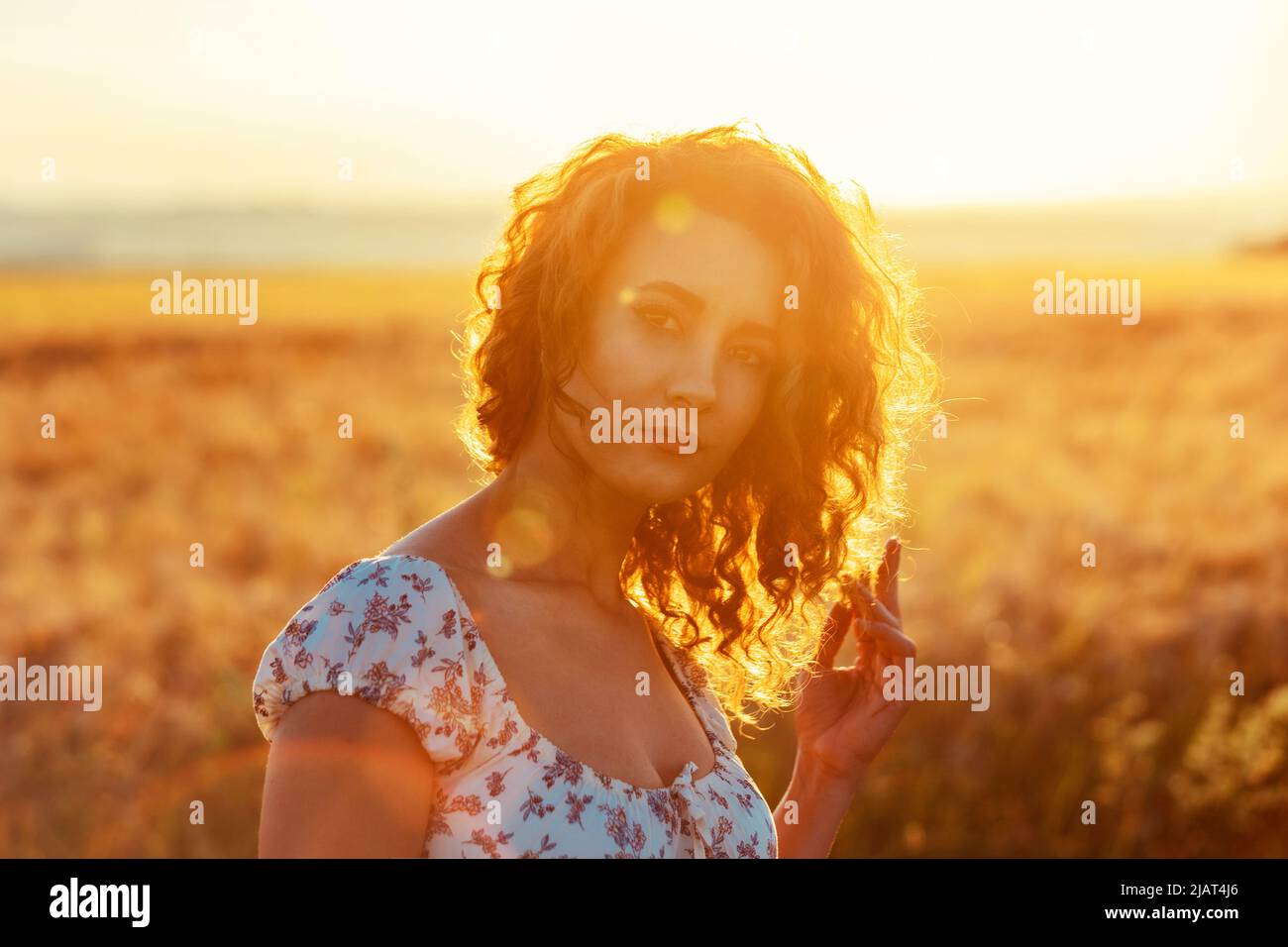 young moroccan woman, with brown curly hair, standing in a wheat field, while the sun ist setting in the background and blinding the camera Stock Photo