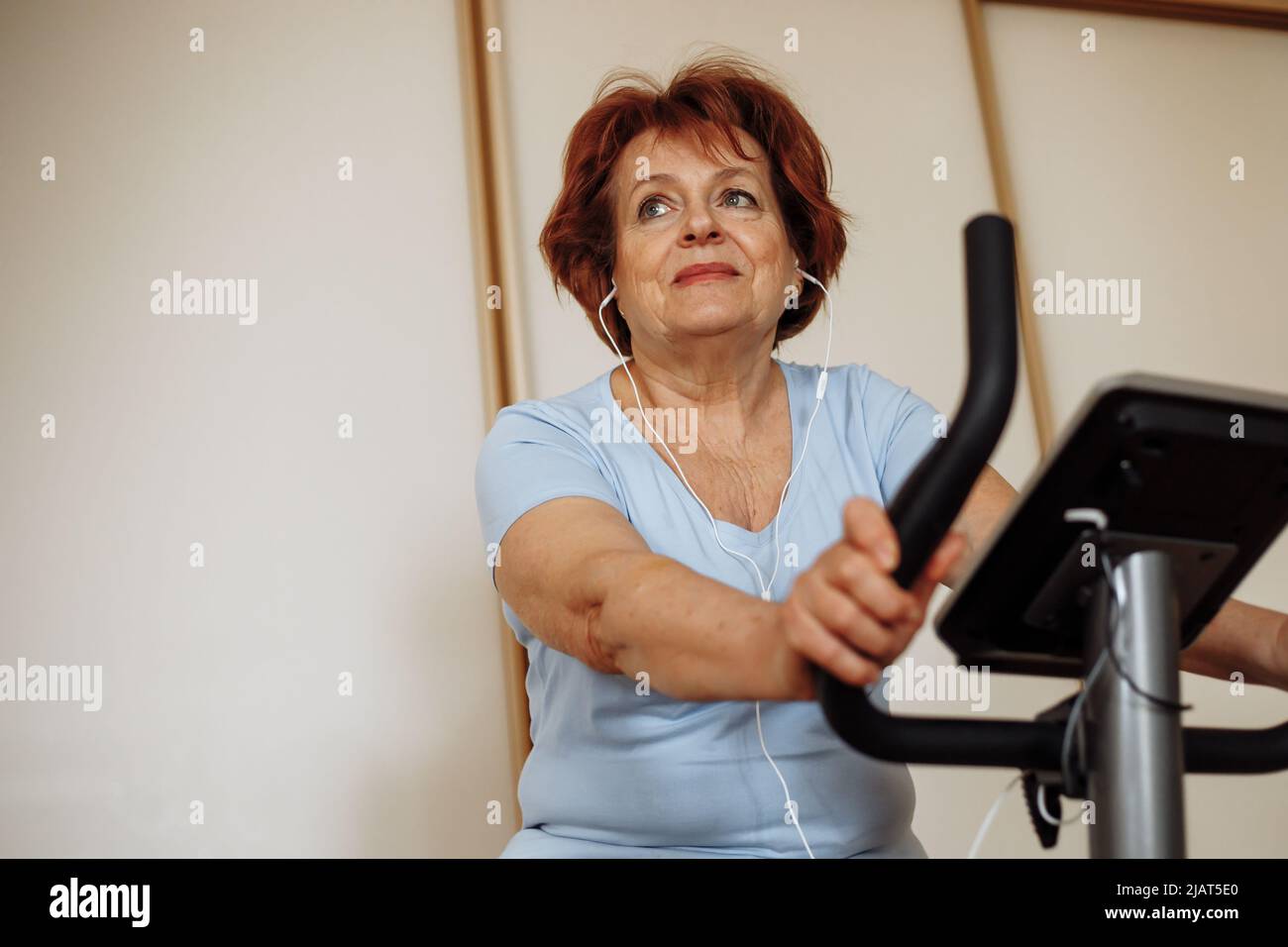 Portrait of elderly woman with dark hair, make-up wearing blue T-shirt, sitting on stationary bicycle, doing exercises. Stock Photo