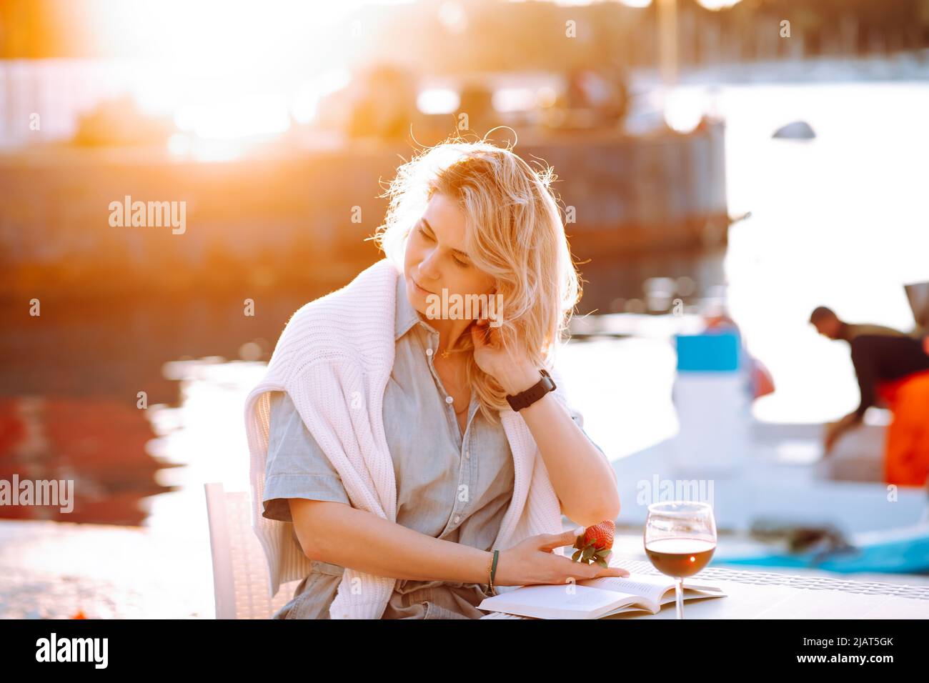 Portrait of young beautiful woman with long wavy fair hair sitting at table with glass of red wine and book at sunset. Stock Photo