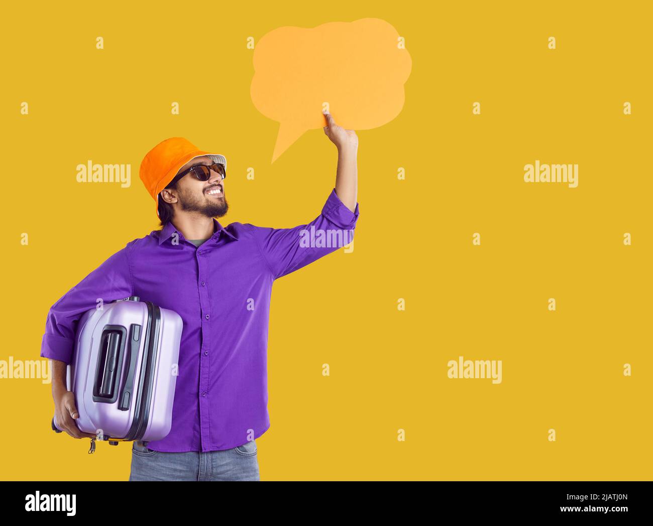 Happy traveler standing on orange background, holding suitcase and showing speech bubble Stock Photo