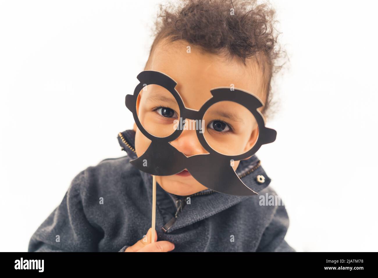 medium close up shot over white background of an interracial cute toddler baby boy holding black fake paper moustache and glasses. happy childhood concept. High quality photo Stock Photo