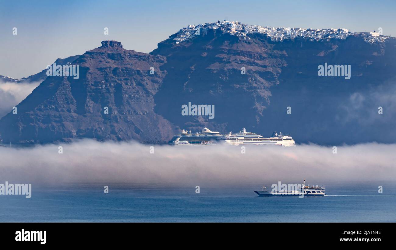 An early morning cloud lying on the water of Santorini's caldera partly obscures a cruise ship that is passed by a ferry boat moving in the sunlight Stock Photo