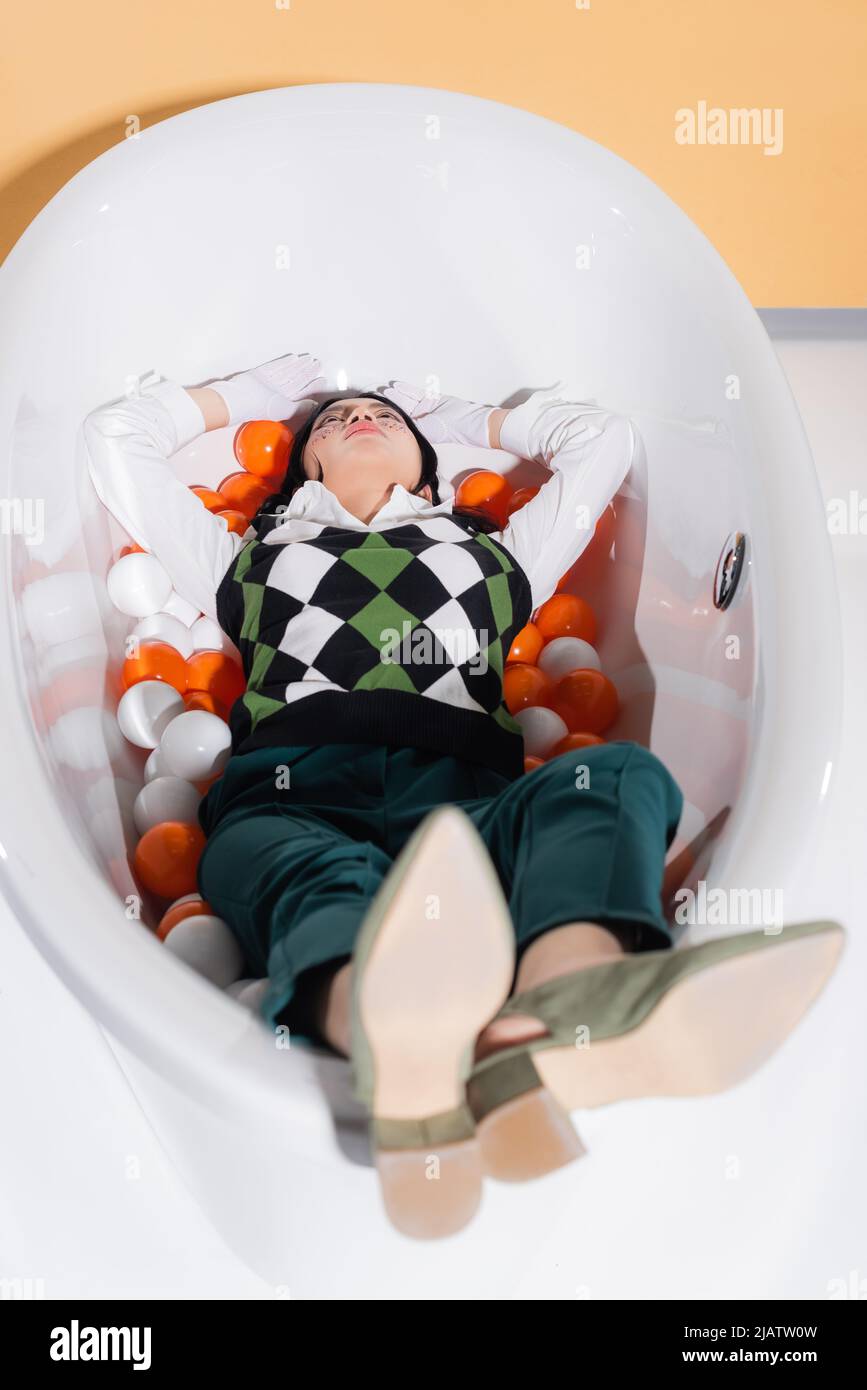 Stylish asian model in gloves lying in bathtub with balls on orange