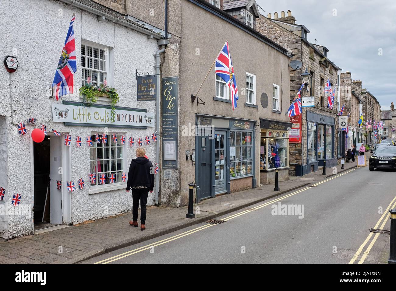 A street in a pretty tourist town on the edge of the Yorkshire Dales and Lake District showing storefronts and a shopper with the Union Flag flying in Stock Photo