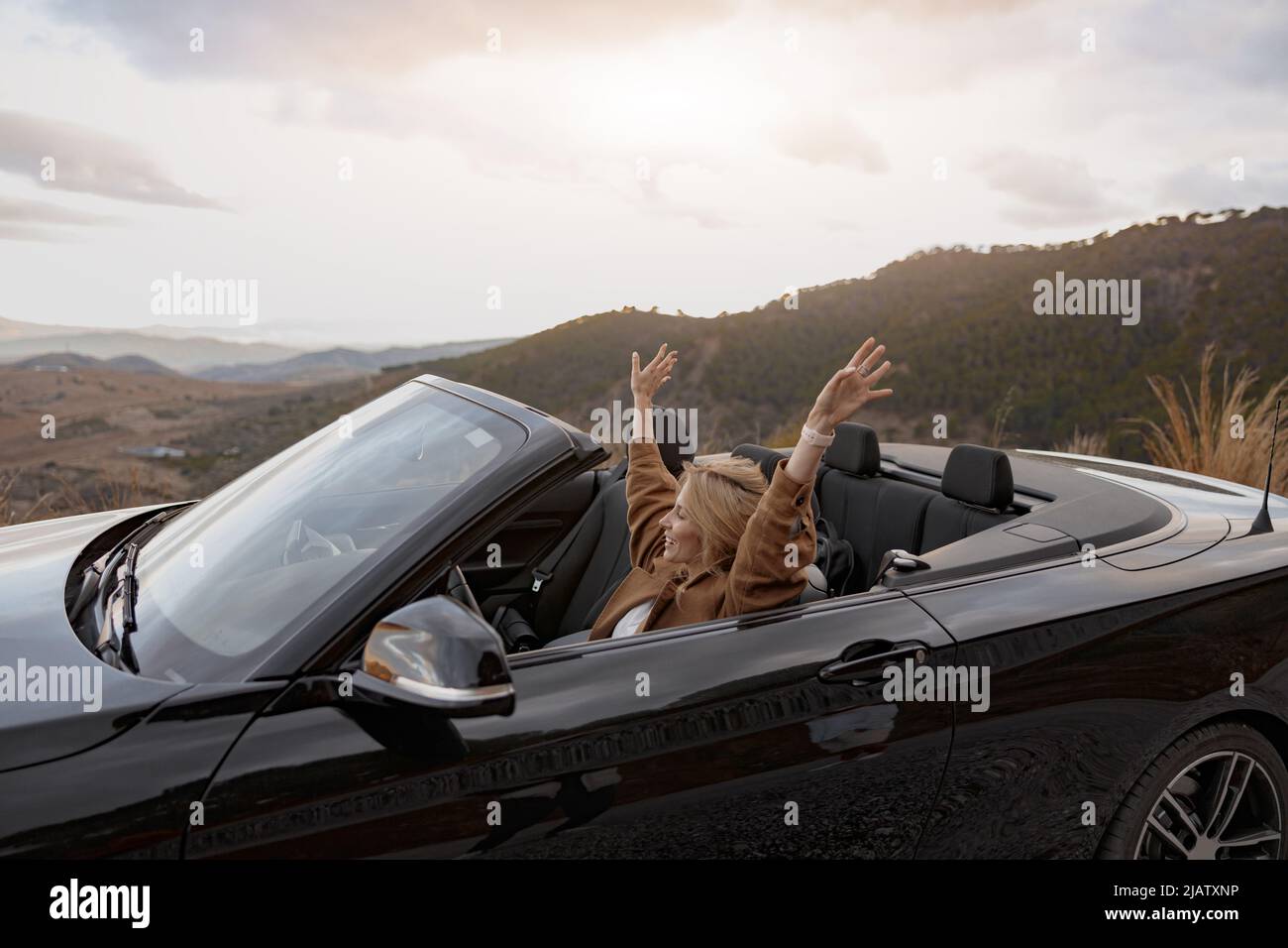 Young happy smiling female traveling in the convertible car by mountainside, side view Stock Photo