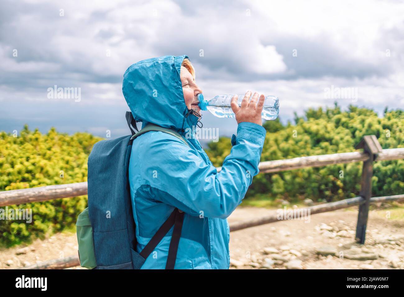 Tired female 50s hiker drinking water from the bottle with backpack