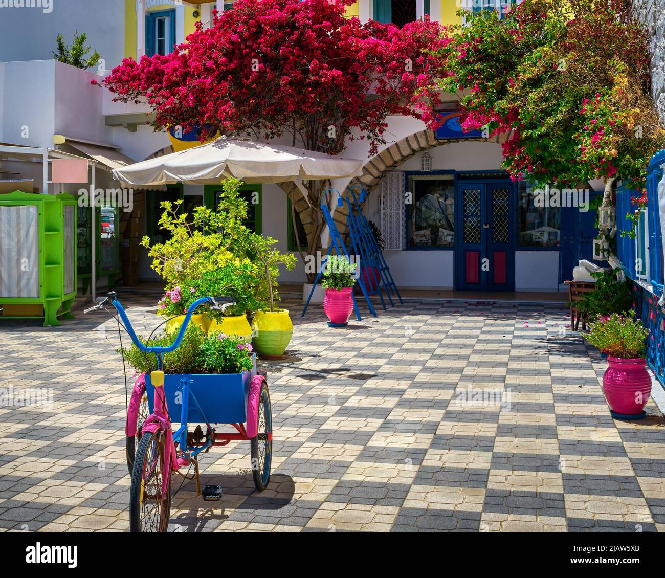 Beautiful corner shop in Adamas town, Milos, Cyclades islands. Greece. Summer sunshine, bougainvillea, flower pots, vines, cute colorful bicycle,  Stock Photo