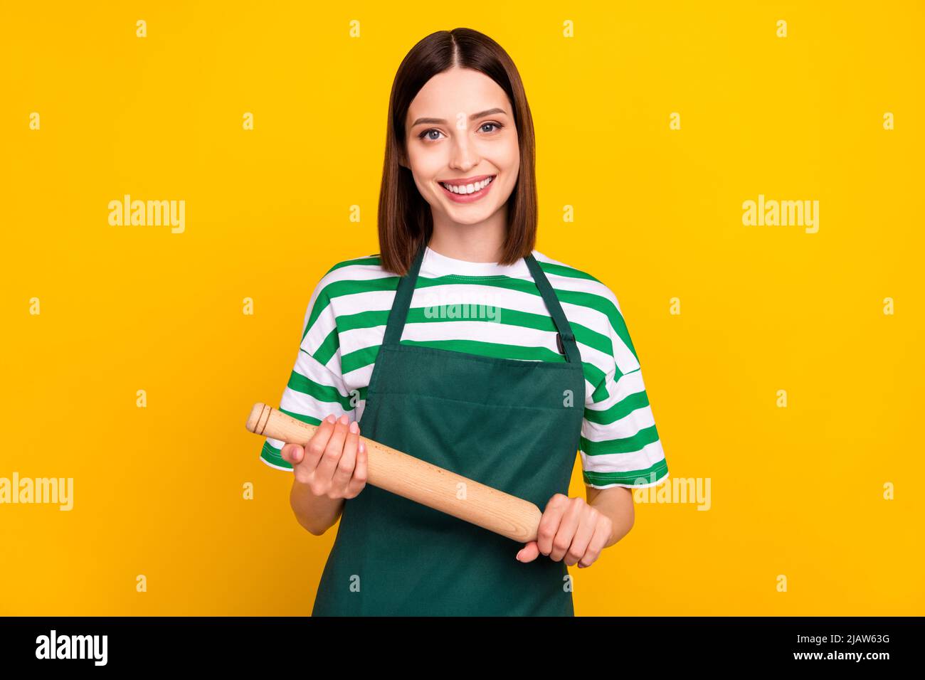Photo of optimistic brunette young cooker lady hold rolling pin wear striped t-shirt apron isolated on yellow color background Stock Photo