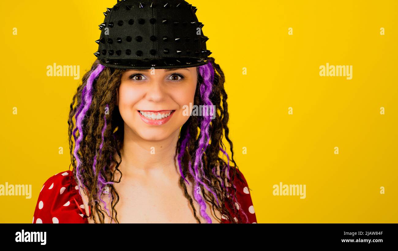 Young handsome stylish feman in black cap on yellow background. Hipster girl looking at camera with dreadlocks. Stock Photo