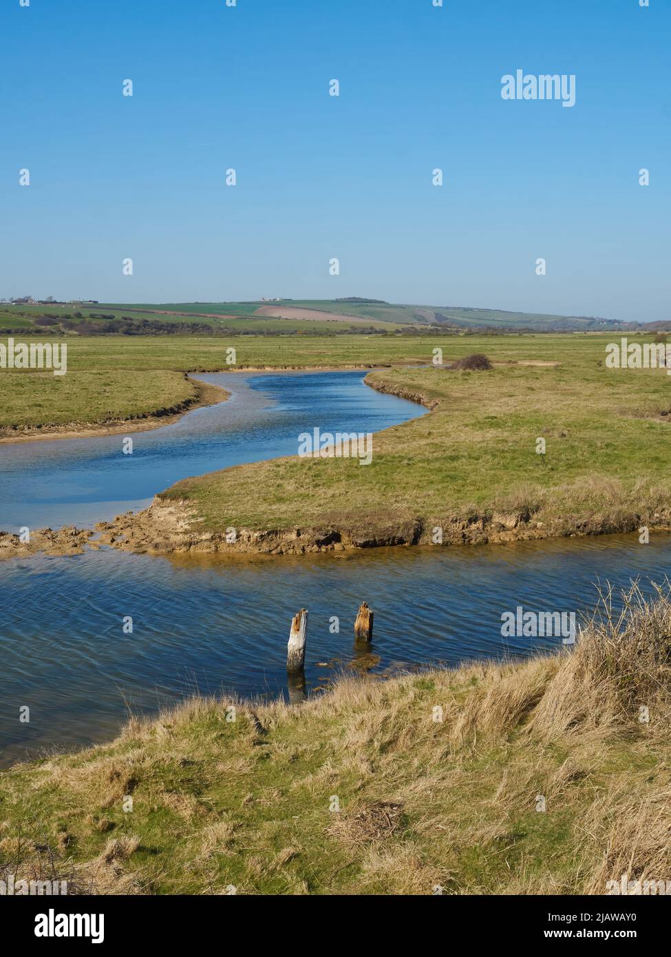 A sunlit river meandering through the open fields of a wetland area with rolling hills on the horizon and a summery blue sky overhead. Stock Photo