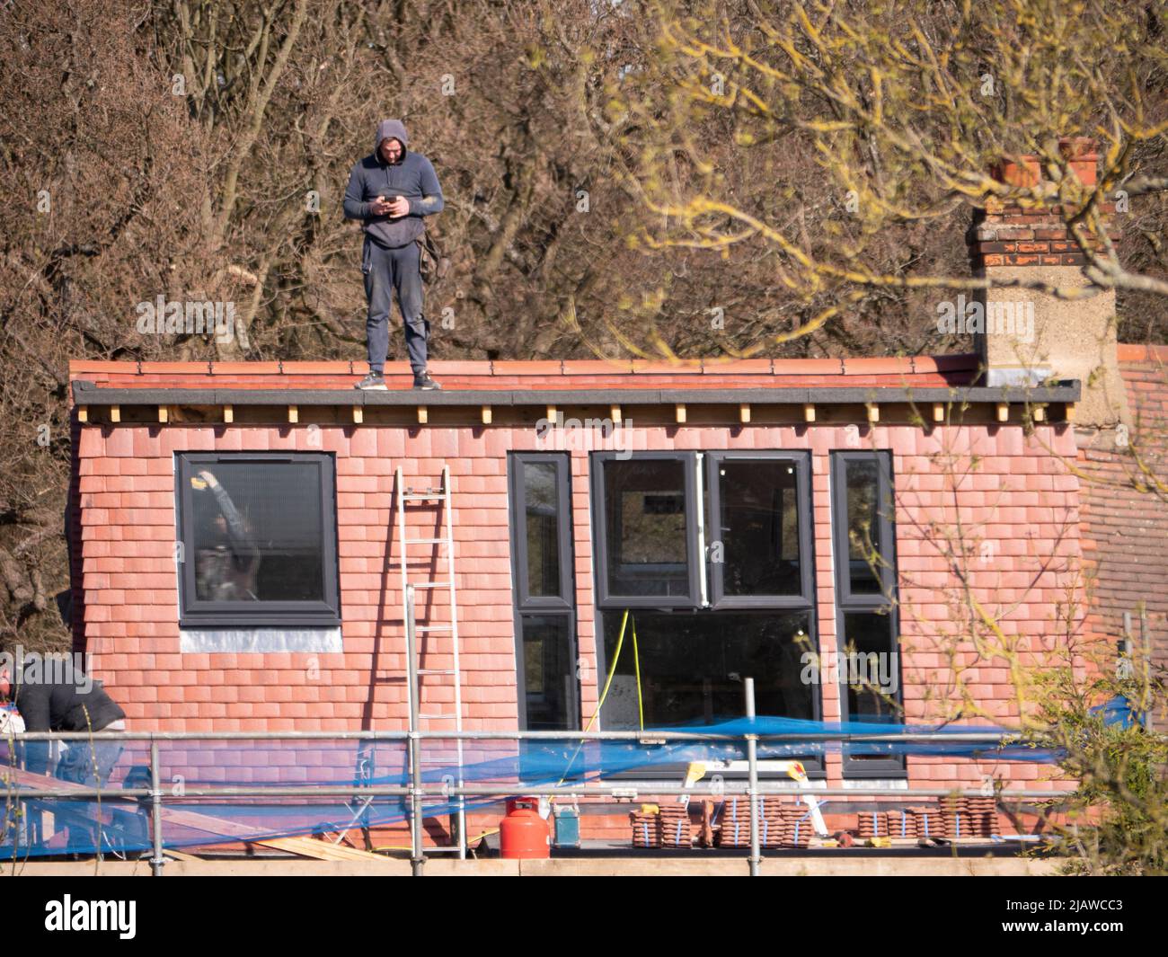 Roofer standing on roof of loft extension with mobile phone near Epping Forest, some areas have poor coverage of phone signal especially in some rural areas. Stock Photo