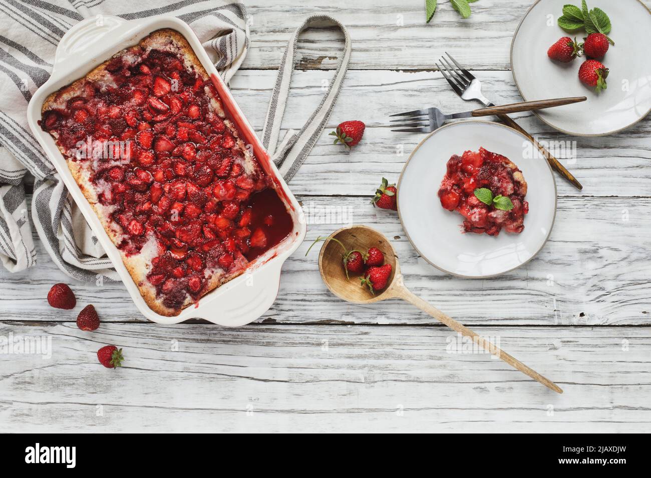 High angle top view of sweet homemade strawberry cobbler or Sonker baked in a red ceramic pan with serving in saucer, apron and wooden spoon. Stock Photo